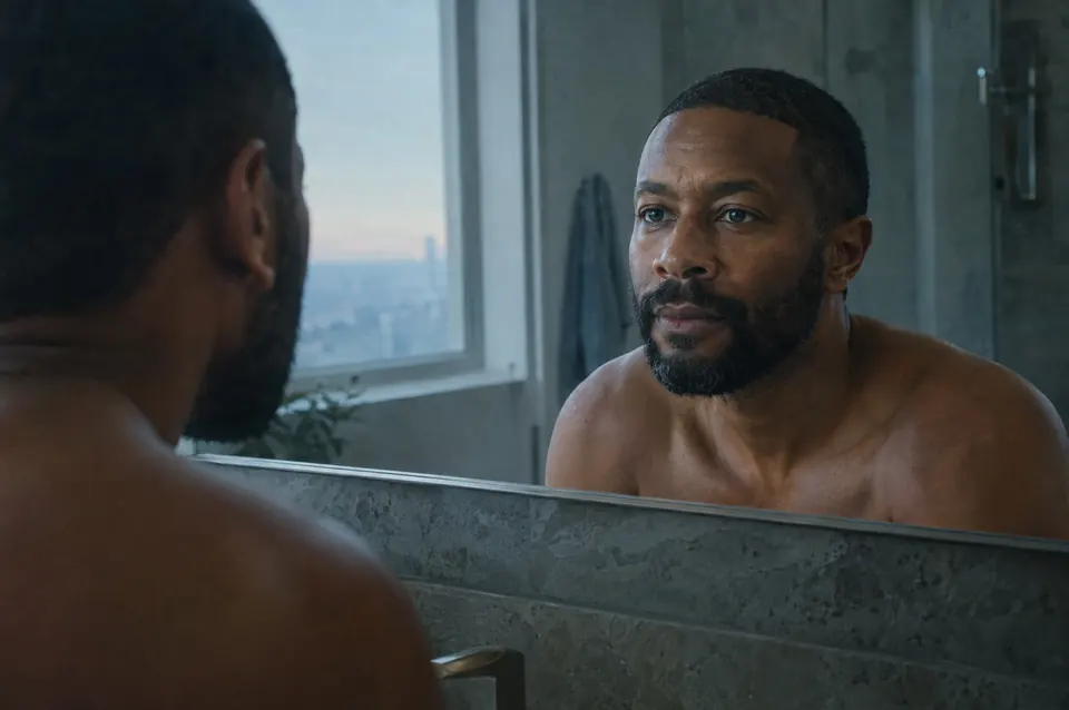 A man examines his reflection in a hotel bathroom mirror in early morning light, the city faint through the window.