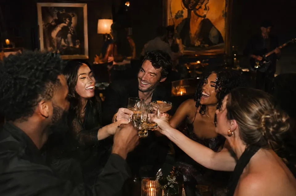 A group of friends raise glasses together in a dark, low-lit lounge.