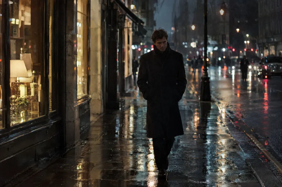A man in a dark overcoat walks a rain-wet city street at night, shop windows glowing.