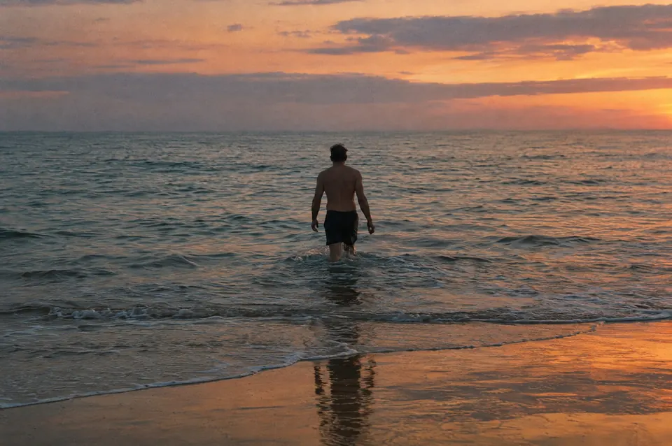 A man wades into a calm sea at sunset, the sky orange over still water.