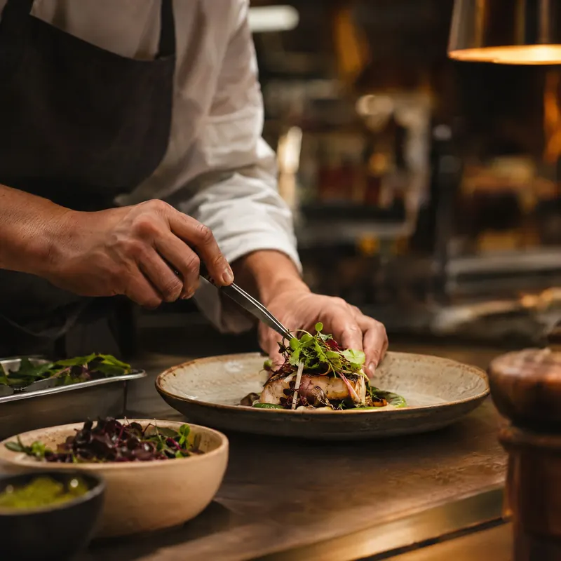 A chef’s hands plate a dish of greens and fish on a warm, dark kitchen counter.