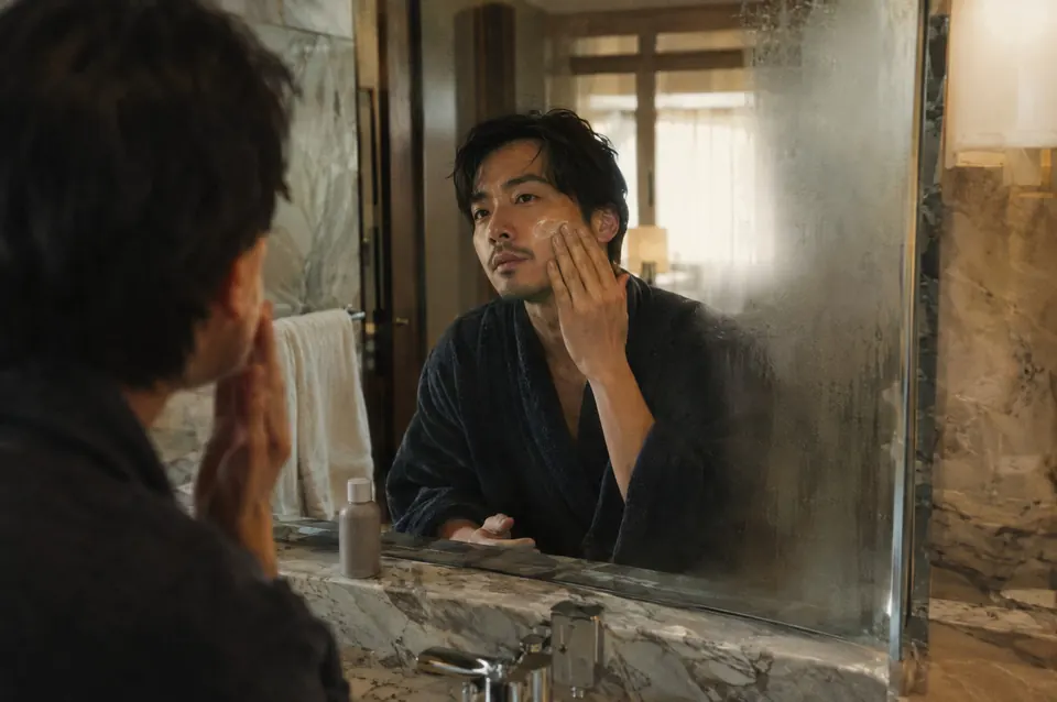 A man applies moisturizer in a steamed bathroom mirror, framed by pale stone.