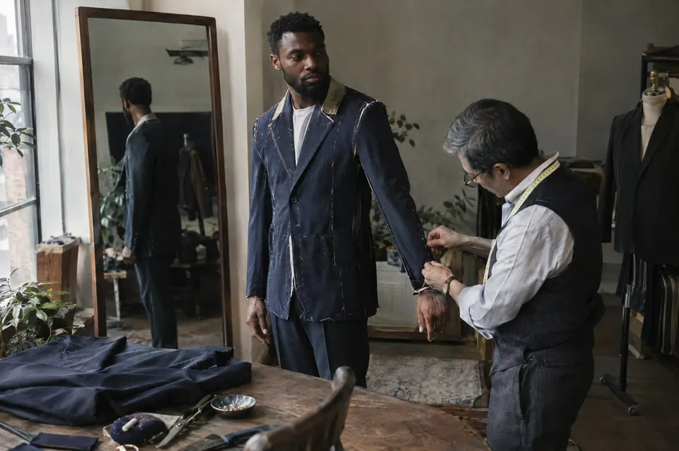 A tailor adjusts the sleeve of a navy jacket on a man standing before a full-length mirror in a bright fitting room.