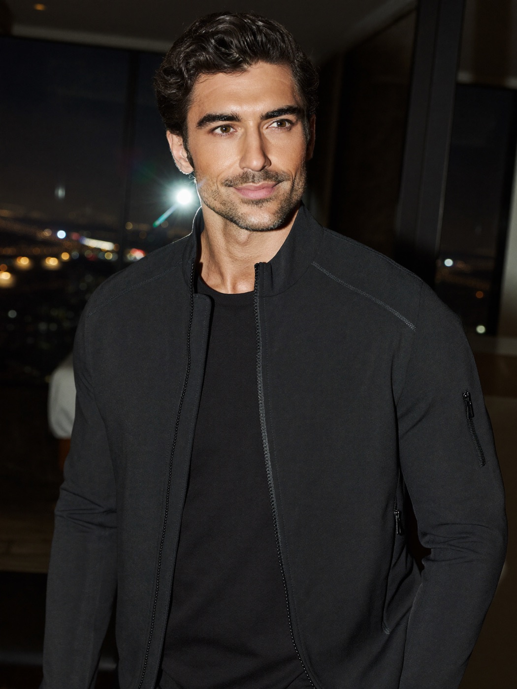 JAX, EYEGASMIC AI coach for travel fitness, in a black travel jacket by a hotel window at night