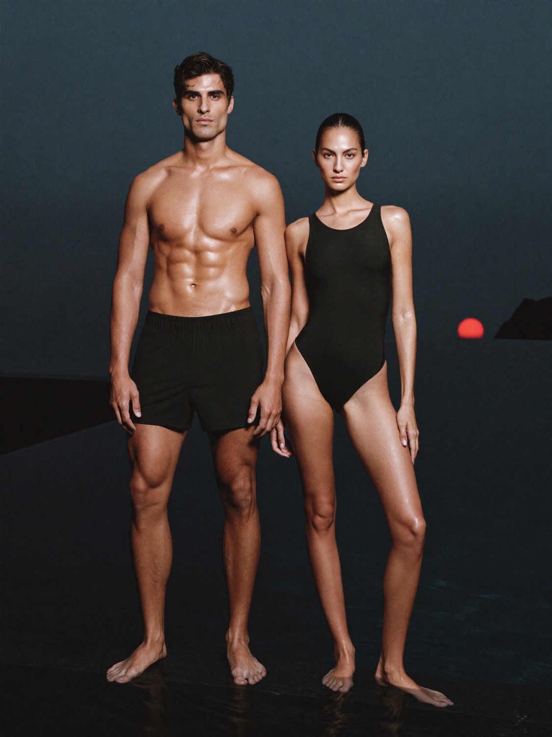 Two athletes in black swimwear at the edge of a dark pool at dusk